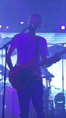 The video shows a band performing on stage. A male guitarist wearing a white t-shirt and jeans is in the foreground, playing a guitar. He is illuminated by bright stage lights. Behind him, other band members are visible, including a drummer. The stage is decorated with a backdrop displaying an abstract image, and various speakers and amplifiers are set up. The audience is partially visible at the bottom of the frame, with some individuals holding up their phones to record the performance. The lighting is predominantly purple and white, creating a concert atmosphere.