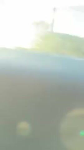 The video shows a blue four-door car with four men inside. The driver is wearing a white hat. The car is driving on a street with power lines visible in the background. The sun is shining brightly. The camera is still and focused on the car.