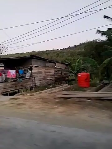 The video shows a side view of a modest roadside dwelling with laundry hanging on a line. In front of the dwelling, there are several wooden stalls or tables. A national flag, possibly red and white, is visible. A blue and white flag is also present. The dwelling appears to be constructed from wood and is situated next to a road. Further down the road, a motorcycle is parked in front of a building with a sign that reads "NAROK BLACK". A person wearing a backpack and shorts is walking away from the camera towards the building. The environment appears to be rural or semi-rural, with trees and a hillside visible in the background. The sky is overcast. The video is shaky, suggesting it was filmed from a moving vehicle.