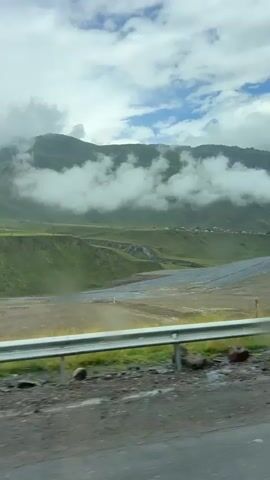 The video shows a scenic view from a moving vehicle. The landscape consists of rolling green hills and mountains partially covered in clouds. In the distance, a small village with scattered houses is visible on a plateau. A wide, flat area, possibly a riverbed or agricultural land, lies in the foreground. A metal guardrail runs along the side of the road. There are no people visible in the video, only natural scenery and man-made structures like the guardrail and a distant electrical tower. The sky is overcast with visible clouds. The overall atmosphere is peaceful and natural.