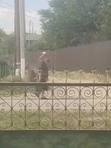 A man in a red shirt and white hat walks with a cane. A woman in a dark dress and headscarf walks in front of him. They are on a sidewalk next to a fence and a brown fence. Trees and houses are visible in the background.