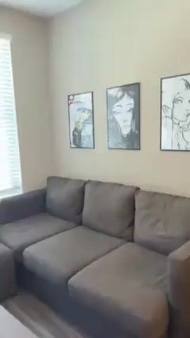 The video shows a corner of a living room with a gray sectional sofa and two framed pictures hanging on the wall. A skateboard is mounted vertically near a window with white blinds. The camera moves to focus on the view from the window, revealing a courtyard with a swimming pool surrounded by lounge chairs and people sunbathing. The surrounding buildings are painted in shades of blue and gray.