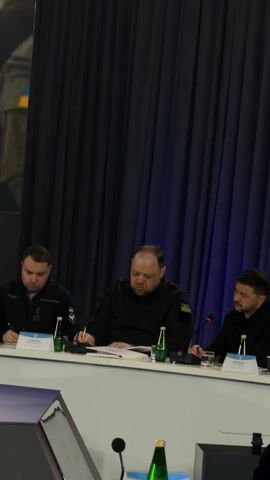 Three men are seated at a white table, participating in what appears to be a formal indoor meeting in Barvinok, Ukraine. All three individuals are wearing dark, professional attire, possibly a uniform.

On the right, Ukrainian President Volodymyr Zelenskyy, identified by his name placard as "ЗЕЛЕНСЬКИЙ В." (Zelenskyy V.), has a short beard and is shown listening attentively, with his chin resting on his hand. In the center, a heavier-set man with a beard is actively speaking, gesturing with his right hand while holding a pen in his left. A yellow and blue patch, likely representing the Ukrainian flag, is visible on his left sleeve. His name placard is partially obscured, but the visible letters strongly suggest "КОСТІН" (Kostin). On the left, a third man, identified by his name placard as "СТЕФАНЧУК" (Stefanchuk), is focused on writing on a document placed before him.

The table is equipped with microphones, green glass bottles of water, drinking glasses, and stacks of paper. Dark blue and black curtains serve as the backdrop for the meeting. The expressions on the faces of the participants convey seriousness and concentration.