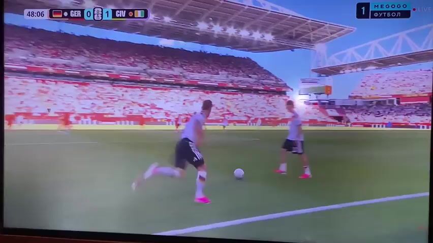 The video shows a soccer game being played in a stadium. The field is green grass, and there are white lines marking the boundaries. There are two teams of players, one in orange jerseys and the other in white and black jerseys. The crowd in the stands is mostly wearing white. A soccer ball is visible on the field. The scoreboard at the top of the screen indicates the game is between Germany and another team, with the score at 1-0. The time elapsed in the game is 48:06. The camera is focused on the action on the field, with a slight tilt upwards. The stadium appears to be modern, with a tiered seating arrangement and a roof structure. There are advertising boards around the stadium. The overall lighting suggests it is daytime.