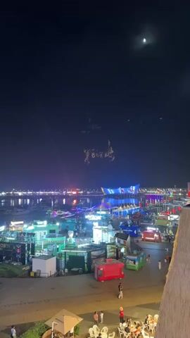 The video shows a night scene from a raised vantage point overlooking a bustling waterfront area. In the foreground, there are several tables and chairs with people dining outdoors. Beyond them, the waterfront is filled with numerous small stalls or kiosks, many of which are illuminated with bright, colorful lights. There are boats docked in the water, also adorned with lights. In the distance, a large, modern building with striking blue and white lights is visible, reflecting on the water. A bright beam of light, possibly from a searchlight, shines upwards from the waterfront area into the dark night sky. The moon is also visible in the upper right corner of the frame. At one point, what appear to be fireworks or a light display forming characters in the sky can be seen. People are walking around the waterfront area, and some are interacting with the stalls. The overall atmosphere is lively and festive, suggesting a night market or a public event.