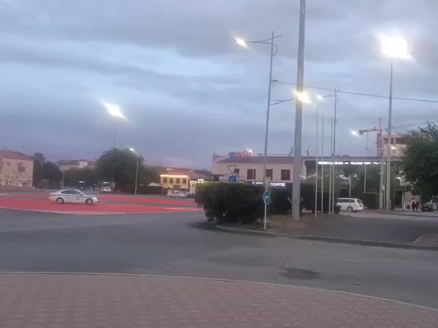The video shows a street scene at dusk. There are streetlights on and the sky is cloudy. In the foreground, a red-painted crosswalk is visible. On the left side of the screen, a police car with flashing blue lights is stopped at the intersection. A white car is driving past the police car. In the background, there are several buildings, including what appears to be a commercial establishment with the word "COFFEE" visible. There is also a construction crane in the distance. The overall impression is of a busy intersection during an incident or a police operation.