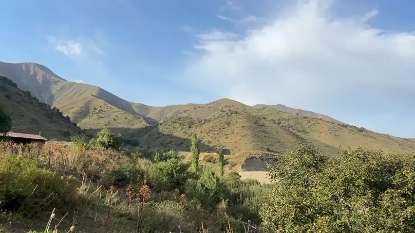 A panoramic video pans across a mountainous landscape. The scene is dominated by rolling hills covered in dry grass and scattered bushes, with a few trees visible. The sky is partly cloudy with patches of blue. In the foreground, there are more dense bushes and dry vegetation. A small building with a dark roof is visible on the left side of the frame. A dirt embankment runs horizontally across the middle of the scene, with more vegetation and some white structures, possibly small houses or tents, visible further in the distance on the top of the embankment. The overall impression is one of a rural, natural environment under a bright sky.