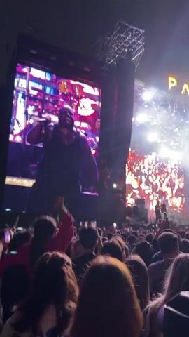 The video shows a concert performance with a large crowd in front of a stage. On the main screen behind the stage, a male performer in a dark vest is singing into a microphone. To the right of the main screen, another screen displays abstract colorful visuals. Above the stage, the words "PARK LIVE" are illuminated in gold lettering. The audience is holding up cell phones to record the performance, and some are waving their hands. Smoke effects are present on stage, and bright stage lights illuminate the scene. The overall atmosphere appears to be energetic and celebratory.