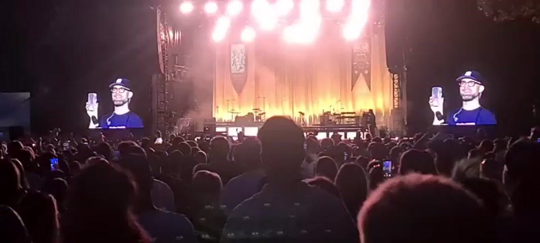 The video shows a live concert from the perspective of the audience. A large stage with bright lights and a backdrop is visible in the distance. On either side of the stage, large screens display a live feed of a man wearing a baseball cap, glasses, and a blue t-shirt, holding up a glass. In the foreground, the backs of many audience members are visible, most of whom are holding up their phones, presumably to record or take pictures of the event. The overall atmosphere appears to be one of excitement and engagement.