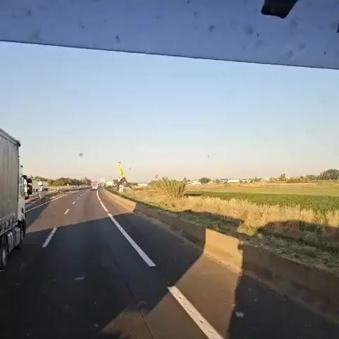The video shows a view from a vehicle driving on a highway under a clear, blue sky. The highway has multiple lanes with white lane markings and a concrete median barrier on the right side. Adjacent to the highway, on the right, there's a grassy area leading to a field with some vegetation. 

The most striking feature is a large, stylized sculpture of a person made of yellow and black geometric shapes. The figure appears to be walking, with a briefcase in hand and a box-shaped head.

Further into the background, there are hints of buildings and other structures in the distance. The scene suggests a rural or suburban area near a major transportation route.