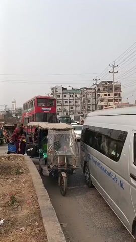 The video shows a bustling street scene in what appears to be a South Asian city.  The road is packed with a variety of vehicles including several auto rickshaws, a red double-decker bus, a grey van with "Incepta Pharma" printed on it, other buses, cars, and a cart being pulled by a person.  Several people can be seen walking or standing along the edge of the road. The sky is overcast. Buildings are visible in the background.  Power lines are strung overhead.