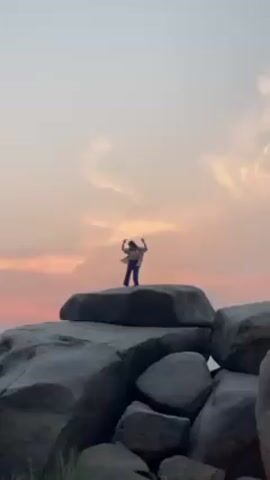 A person is standing on a large rock formation against a sky with pink clouds. The person is wearing a white top and blue pants. They raise their arms in the air and then spin around. The rocks are gray and irregularly shaped. The sky is a mix of pale pink and light gray. There is some green grass at the base of the rocks.
