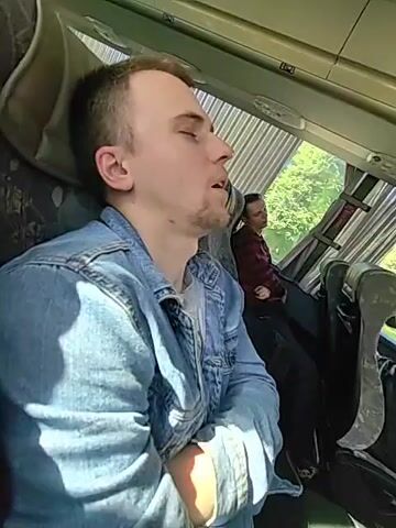 This is a short video clip showing a man asleep on a bus, with his mouth open. He is wearing a denim jacket over a gray t-shirt. His eyes are closed and his head is tilted back. In the background, another passenger is visible, also appearing to be dozing. The bus appears to be traveling, as indicated by the blur of trees and buildings outside the window. The light suggests it is daytime, likely late morning or early afternoon, with bright sunlight filtering through the window. The setting is the interior of a bus, with patterned upholstery on the seats and blinds covering the windows. The overall mood is one of peaceful slumber, perhaps a consequence of a long journey. There is no visible text in the media file.