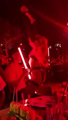 Here is a description of the video:

The video is shot in a dark, red-lit nightclub setting. A woman with long blonde hair pulled into a high ponytail is the focus. She is wearing a revealing outfit consisting of a black harness-style top and bottom set, accessorized with thigh straps. She's interacting with a seated male patron. She appears to be performing or dancing in front of him, holding what looks like a glowing red light bar. The man is wearing a light-colored shirt and has his hands raised as if participating in the performance.

In the foreground, there's a table with plates and skewers of food. Other people can be seen blurred in the background, indicating a busy nightclub environment. The overall atmosphere is one of entertainment and nightlife.