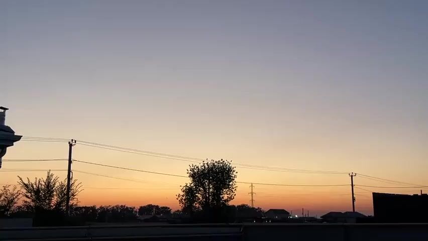 The video shows a dusky sky with an orange glow near the horizon. Silhouetted against the sky are power lines, utility poles, trees, and the outlines of houses. The scene appears to be a rural or suburban setting at either dawn or dusk. There are no people visible in the frame.
