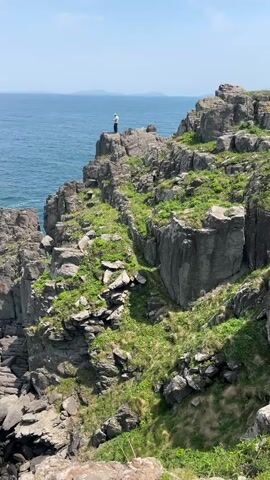 The scene is a rocky coastline with a person standing on a cliff's edge. The individual is facing away from the camera, towards the ocean, with their arm raised slightly. The cliffs are composed of dark gray rocks, interspersed with patches of green vegetation. The ocean is a deep blue, stretching to the horizon under a light blue sky with a few clouds. Waves can be seen crashing against the base of the cliffs. The camera's perspective is elevated, looking down at the scene.