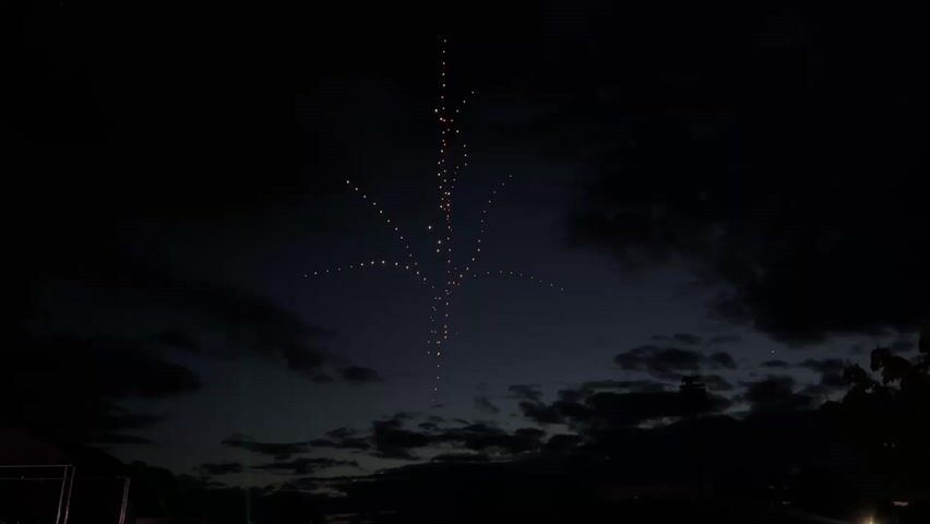 The video shows a dark, almost black, sky with scattered clouds and a strange light formation that looks like a tree. The "tree" is made of many small, faint lights arranged in a trunk-like vertical line with branches extending from it. The branches also consist of small, faint lights. Some barely visible trees are silhouetted against the bottom of the shot.