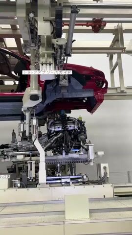 A robotic arm is installing a shock absorber onto a car engine assembly. The engine assembly is suspended from above. The car body is partially visible at the top of the frame. Text overlays read "ショックアブソーバガイド" which translates to "Shock Absorber Guide". The environment appears to be a factory floor.