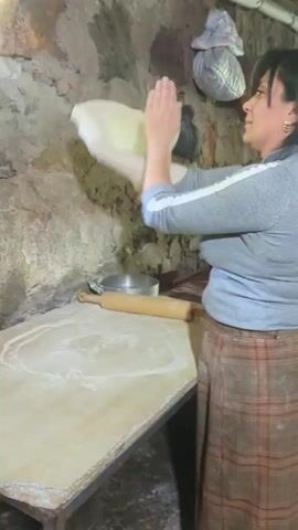 A woman is shown stretching a large piece of dough. She is wearing a gray sweatshirt, a plaid apron, and has dark hair. The dough is light in color and appears to be very thin and pliable. The woman is standing in front of a wooden table, which is dusted with flour. A rolling pin is visible on the table. The background suggests a rustic setting, possibly a kitchen or workshop, with a stone wall and some items hanging on it. The lighting is dim, and there's a window in the background, allowing some natural light to enter. The woman is looking at the dough and appears focused on her task.