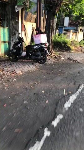 The video shows a street scene. In the foreground, a motorcycle is parked next to a sidewalk. A person wearing a hijab and carrying a pink cooler bag is standing next to the motorcycle. In the background, there is a building with a tiled roof and a tree. Further down the street, another person wearing a helmet and a backpack is walking on the sidewalk. The environment appears to be an urban or suburban area with a paved road and a sidewalk. The lighting suggests it might be daytime, possibly early morning or late afternoon due to the long shadows.