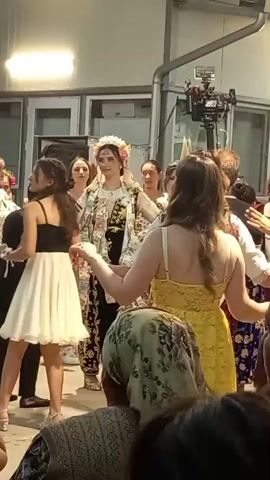A group of people are gathered around a young woman dressed in traditional clothing and a floral headdress. The woman appears to be the center of attention, with others facing her and some reaching out. A video camera on a tripod is positioned behind the group, suggesting the event is being filmed. The setting appears to be an outdoor or semi-outdoor space with a building in the background. Some individuals are wearing festive attire, and the overall atmosphere seems celebratory. The music playing in the background is a lively tune.