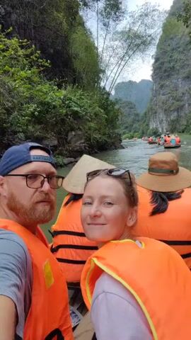 A man and a woman wearing orange life vests are taking a selfie on a boat. In the background, several other boats filled with people can be seen traveling up a river in a mountainous, lush environment. The woman has sunglasses on her head and is smiling for the camera. The man is wearing glasses and a baseball cap and has a beard. Behind them on the boat are two people wearing traditional conical hats. The river is calm, and the weather appears overcast.