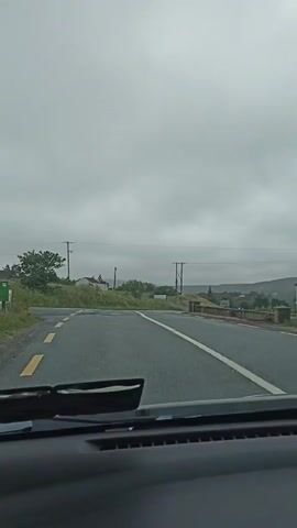 The video shows a view from inside a vehicle driving on a wet road. The sky is overcast and gray. The road curves to the right, with a yellow line on the left and white lines on the right. There are utility poles and some vegetation along the sides of the road. In the distance, there are hills and some buildings. The dashboard of the vehicle is visible in the foreground. The windshield wipers are visible and appear to be stationary.
