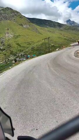 The video shows a first-person perspective from a motorcycle, driving on a winding mountain road. The road is paved with asphalt and has tire tracks visible. On the left side of the road, there is a steep, green, grassy mountain slope with scattered rocks and a few trees. Further down the slope, a small village or settlement with houses and a church can be seen. Power lines with wooden poles run alongside the road. On the right side of the road, the mountain is also steep, with more rocks and sparse vegetation. In the distance, majestic snow-capped mountains rise against a bright blue sky with scattered white clouds. Other motorcycles are visible on the road ahead, indicating that this is a scenic route. The camera appears to be mounted on the handlebars of the motorcycle, providing a dynamic view of the ride. The overall impression is one of adventure and exploration in a beautiful, rugged natural landscape.