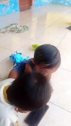 The video shows two young children on a tiled floor. The child in the foreground is wearing a white shirt with yellow accents and is looking down at a smartphone. The second child, slightly further away, is lying on their stomach wearing a blue shirt and dark bottoms, with a blue toy behind them. In the background, a bottle of white liquid and some folded fabric are visible on the tiled floor near a wall with a window and a door. The second child then sits up and puts on a shoe.