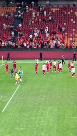 In the video, a soccer team dressed in red and white uniforms is on the field near the sidelines. One person in a neon green vest and another in a yellow jacket stand near the team. The players acknowledge a crowd of spectators in stadium seating that is primarily red. Many people in the crowd are standing and raising their arms. Some are holding red flags. The camera movement is shaky.