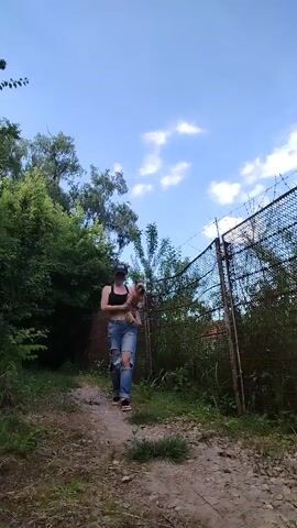 A woman with a dark cap, black bra, and ripped blue jeans is holding a tan Yorkshire terrier and walking forward on a dirt path in an outdoor setting. The woman walks slowly towards the camera with the dog in her arms, which she raises a bit. The dog has long hair and is looking in the direction of the camera. The woman smiles and looks at the dog as she holds it. The background features lush green trees and a chain-link fence on the right side. The sky above is clear and blue.