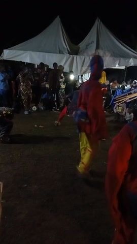 The video shows several people in colorful costumes dancing outdoors at night. The location appears to be a grassy field or yard. Behind the dancers, there is a white tent and a crowd of onlookers, many of whom are holding up phones, presumably to record the performance. The overall scene seems to be part of a cultural event or celebration, with an emphasis on traditional dance and community gathering.