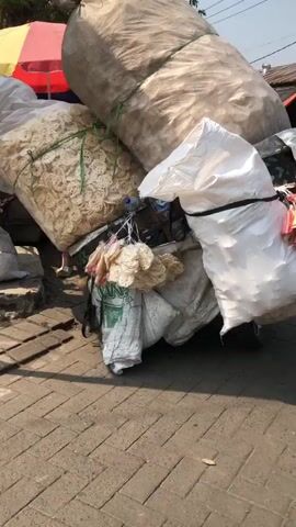 The video shows a street scene with several motorbikes and people. In the foreground, a motorbike is heavily loaded with large sacks and bundles of what appear to be food items. One sack is a very large, light-colored material, and another is white and appears to contain a powdery substance. There are also bundles of packaged items, some with pink and yellow wrappers, and others that look like dried snacks. The motorbike is parked on a brick-paved surface. In the background, other motorbikes are visible, some with riders. One person is seated on a scooter, facing away from the camera, and another person appears to be sitting on a bench under an umbrella. Several stalls or market setups are present, with one displaying white crates filled with red produce, likely onions, and a scale. The environment suggests an outdoor market or street vendor area with trees and buildings in the distance. The lighting indicates it is daytime.