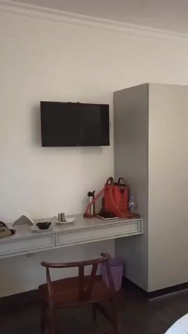 The video shows a view of a hotel room. There is a white wall with a mounted TV above a built-in desk. The desk is cluttered with items like a water bottle, a small red bag, and dishes. A wooden chair is tucked under the desk. Beside the desk is a grey wardrobe with two doors. Next to the wardrobe, a set of shelves are visible. A round wicker basket sits below the shelves. A wooden door is visible further into the room.  A bed is in the foreground, covered in white sheets with a gray runner. Two suitcases are on a shelf at the end of the bed. The floor appears to be dark.  The room seems simple but clean.