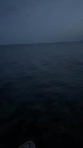 The video begins with a close-up of water, possibly the sea, with a dark sky overhead. The scene then shifts to a person's feet and legs wearing light-colored shoes and dark pants, standing on a dark surface, possibly rocks or soil. The view pans upwards, revealing a dark, wooded area. The video then transitions to a wider shot of a body of water, with a shoreline in the distance. A dramatic sunset is visible on the horizon, with vibrant orange and red hues illuminating the sky above a silhouetted mountain range. The water is choppy, and the sky is filled with dark, dramatic clouds. The overall mood is serene and picturesque.