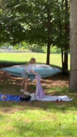 A young woman with blonde hair, wearing a white t-shirt, light-colored shorts, and white sneakers, stands in a grassy outdoor area under trees. Behind her, a light blue hammock is strung between two trees. On the ground in front of her, a blue blanket with white stars and a white and light purple blanket are spread out, with a black bag resting on the blue blanket. She stands upright, adjusts her shorts, and then leans down, looking at the ground and the items laid out in front of her. The background shows a grassy field and additional trees under a bright sky.