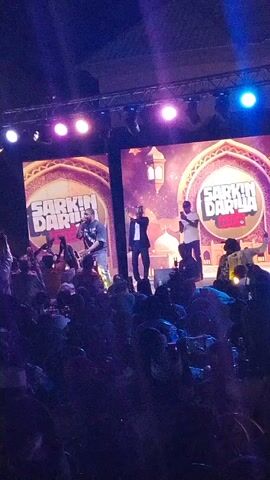 Here's a description of the video:

The video shows a live music performance at an outdoor venue. A crowd is visible in the foreground, many with their hands raised, indicating they are enjoying the show. On stage, three performers are visible under bright stage lighting. Two large video screens flank the stage, each displaying the text "SARKIN DARIA." The performance appears energetic, and the atmosphere seems lively. The event is taking place at night, with the outdoor venue open to the sky.