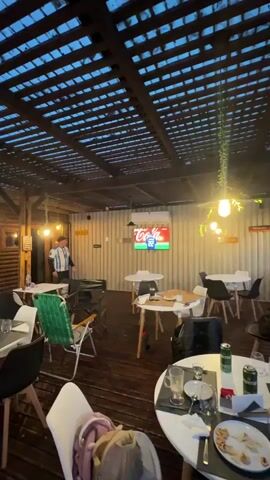The video shows an outdoor cafe at dusk. A man wearing an Argentina soccer jersey walks past a television screen displaying a soccer game. Several tables with chairs are set up in the cafe, some with drinks and food on them. A wooden roof covers the outdoor space, and string lights and hanging plants are visible.