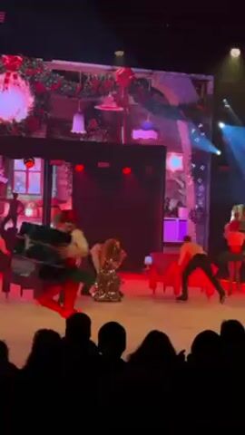 A group of ice skaters perform a Christmas-themed routine in front of an audience. One skater in a red hat, tights, and green tutu carries a large, wrapped gift, while others skate in similar costumes or in black pants and tan shirts. The stage backdrop displays a Christmas scene with wreaths, bows, and a row of houses decorated with lights and stockings. The stage is lit with red spotlights and the ice is a pale, reflective surface. The video is taken from the audience's perspective.