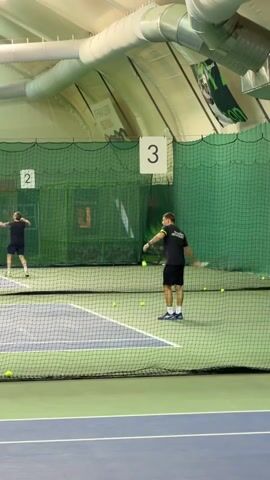 In an indoor tennis court, two men practice tennis. One man, dressed in black shorts and a black t-shirt with yellow trim, hits tennis balls from the right side of the net, near a sign labeled "3". The other man, wearing dark shorts and a gray t-shirt, hits balls from the left side of the net, near a sign labeled "2". There are several loose tennis balls scattered on the ground between the net and the backdrop. The court is blue, and a green net separates the two sides. Above, a corrugated metal ceiling runs horizontally, with a cylindrical air duct and various lights and signage.