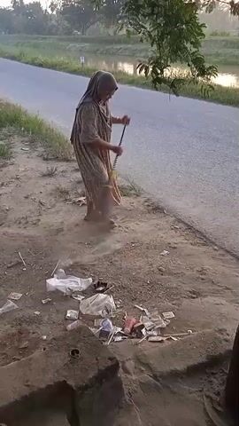 A person wearing a hijab and a long dress sweeps the ground with a broom next to a road. The ground is littered with trash, including plastic bags, wrappers, and cigarette butts. In the background, there is a river and trees. The lighting suggests it is early morning or late evening.