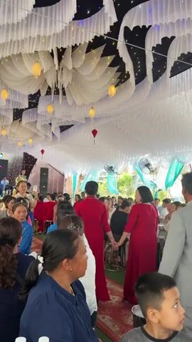 The video captures a festive gathering, likely a wedding reception, taking place under a elaborately decorated canopy. The ceiling is adorned with flowing white fabric drapes, creating a wavy pattern, interspersed with string lights and illuminated yellow and red lanterns. Several ceiling fans are visible, suggesting a warm climate.

Many people are present, both seated at tables and standing, facing towards a central, unseen area. The attendees are predominantly adults, with some women dressed in traditional Vietnamese Ao Dai, particularly in vibrant yellow and red colors. Some individuals are seen walking through the crowd. The overall atmosphere appears celebratory and lively. While the time of day isn't explicitly clear, there's a mix of natural light and indoor illumination. The event is set in Bình An, Vietnam.