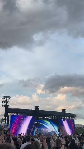 The video shows an outdoor music stage with two large screens displaying images of two singers or performers. In front of the stage, a crowd of people are holding up their phones, presumably to record the performance. The sky above is filled with a mix of gray storm clouds and patches of blue sky. The stage is equipped with lighting and sound equipment. Some palm trees are visible in the far distance.