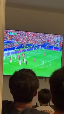The video shows a large television screen displaying a soccer match. The score indicates Portugal 0 and Spain 0. The game is taking place in a stadium filled with spectators wearing red. On the field, players in red and white uniforms are visible, engaged in play near one of the goals. In the foreground, from behind, the heads and shoulders of three people are visible, looking at the television. The lighting suggests an indoor environment, possibly a living room, with the television being the primary light source.