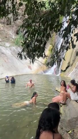 A group of people are at a waterfall. Some are swimming in the pool at the base of the waterfall, while others are sitting on the rocks surrounding the pool. The waterfall is surrounded by lush greenery and rocky cliffs. In the foreground, a woman in a bikini is standing on a rock, looking at the camera. In the background, a waterfall cascades down a rocky cliff face. Several people are visible in the water, either swimming or wading. Others are seated on the bank of the pool. Some vegetation hangs over the water from trees and vines.