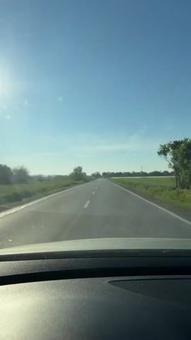 The video shows a view from inside a car driving on a straight, two-lane asphalt road. The sun is visible in the upper left corner, shining in the clear, blue sky. Fields of green vegetation are on either side of the road.  A few trees and utility poles are scattered along the roadside. The car's dashboard is visible in the foreground, providing a view of the road ahead.  The video appears stable, as if the camera is mounted on the dashboard.
