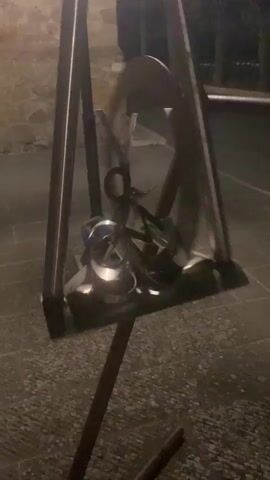 A metallic sculpture consisting of a large circular frame with a smaller circular frame within it. A vertical metal bar passes through the center of both circles. Attached to the vertical bar are several smaller metal pieces that appear to be part of a mechanism. The sculpture is mounted on a dark, textured surface, possibly stone paving. In the background, there is a stone wall and some blurry foliage, suggesting an outdoor setting at night or in low light conditions. The lighting is dim, with some artificial light sources highlighting the sculpture, creating reflections and shadows. There are no people visible in the video. The sculpture is stationary.