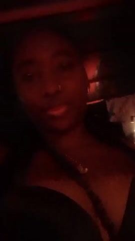 A person is recording a selfie video. The person is a dark-skinned female with braided hair, a nose ring, and a gold necklace. The person is wearing a black top and a black fanny pack with a white object underneath. The background has red and white colors. The person sticks out their tongue.