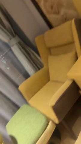 The video captures a slow pan across a cozy seating area, likely indoors in a room or lounge. It begins with a comfortable-looking yellow armchair with a high back and slightly darker brown armrests, accompanied by a light green upholstered footstool or ottoman.

The camera then moves to the right, showing a small, round, light wooden table next to the armchair and footstool. On the table, there is a small white rectangular item with an indistinct red logo or text on it, possibly a room service menu, card, or note.

Behind the furniture, large grey or striped curtains are visible, suggesting a window. Reflections of small, scattered lights, possibly from a city outside, are visible on the windowpane, indicating it is likely evening or nighttime. The overall decor features warm tones, including the yellow of the armchair and table, and a part of a wall with a textured, marble-like pattern.