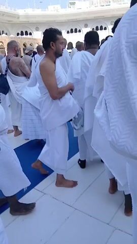 The video shows a group of people, mostly men, walking in the direction of the Kaaba in Mecca, Saudi Arabia. They are wearing white Ihram clothing, which is typically worn during the Islamic pilgrimage of Hajj or Umrah. Some people are barefoot, while others wear simple sandals. The camera follows the group from behind, occasionally showing glimpses of their faces. Some of them carry small black backpacks. The Kaaba, a large cube-shaped structure draped in black cloth, is visible in the distance. The weather appears sunny. The overall impression is one of a religious pilgrimage.